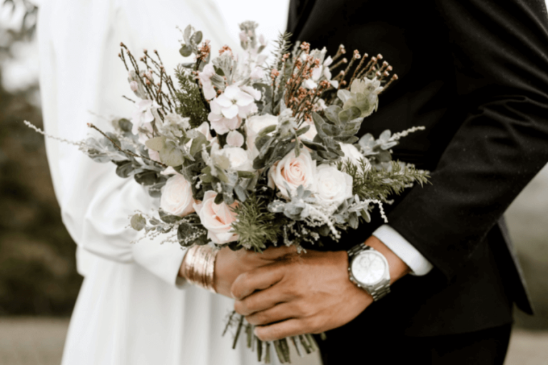 Pareja de novios sosteniendo un ramo de flores, ella vestida de blanco, él en traje negro con reloj plateado.