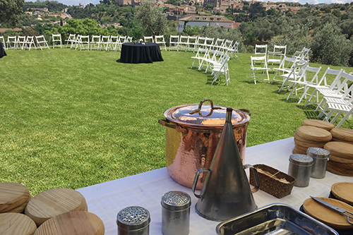 Mesa en un jardín con utensilios para catering. Sillas blancas y mesas negras están dispuestas en el césped.