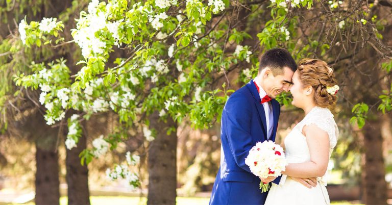 Pareja de recién casados abrazándose bajo árboles en flor, él con traje azul y ella con vestido blanco sosteniendo un ramo.