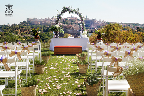 Espacio exterior preparado para una boda, con sillas blancas, flores, un altar y una vista panorámica de una ciudad.