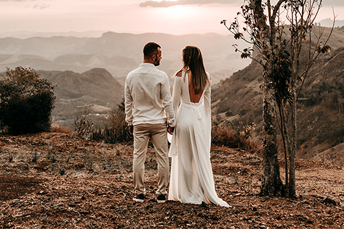 Pareja de espaldas de pie en un paisaje montañoso al atardecer, vestidos de blanco, tomados de la mano.