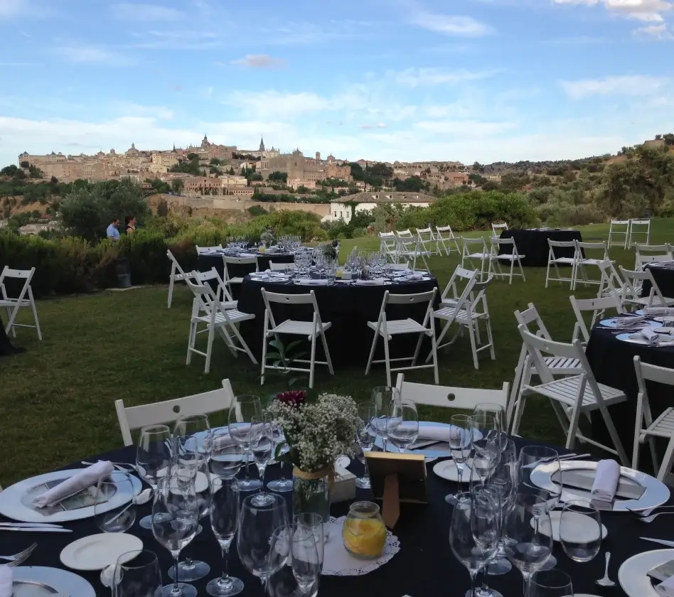 Mesas y sillas blancas preparadas para un evento al aire libre en un campo, con una ciudad histórica al fondo.