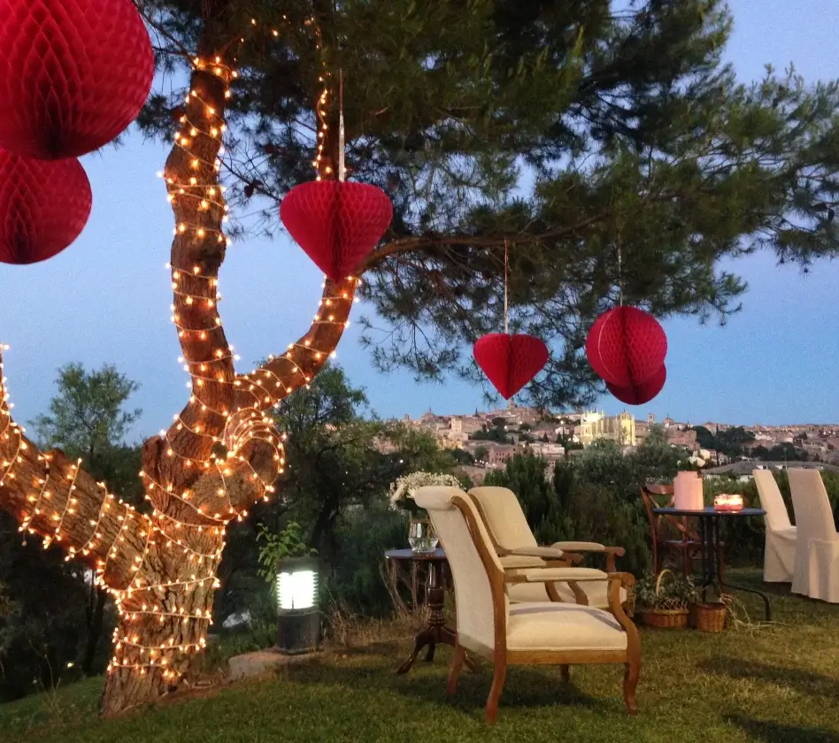 Árbol decorado con luces y corazones rojos, sillas al aire libre y vista de una ciudad al atardecer.