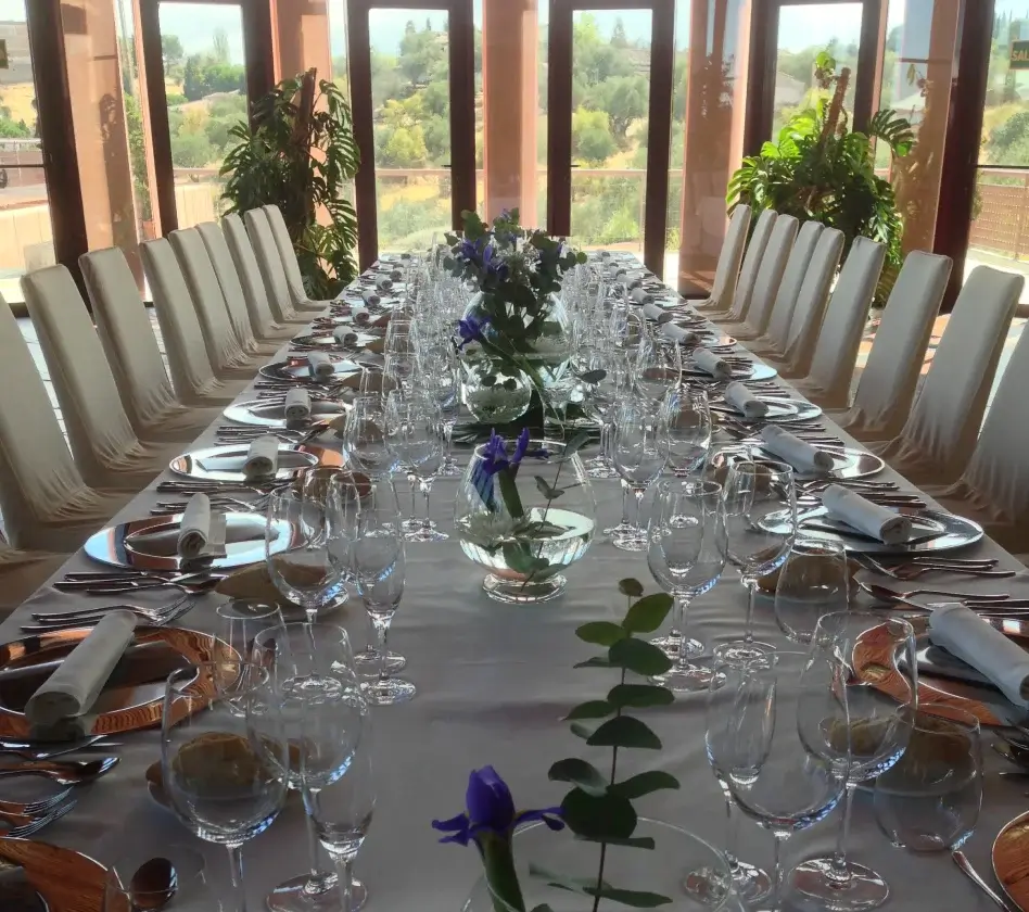 Mesa larga elegantemente preparada con copas, platos y flores azules, rodeada de sillas blancas frente a ventanales.