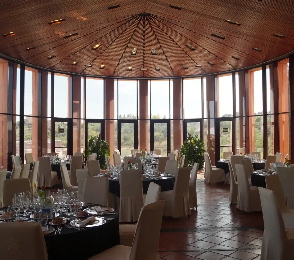 Salón circular con techo de madera, mesas redondas con manteles blancos y sillas, grandes ventanales con vista al exterior.