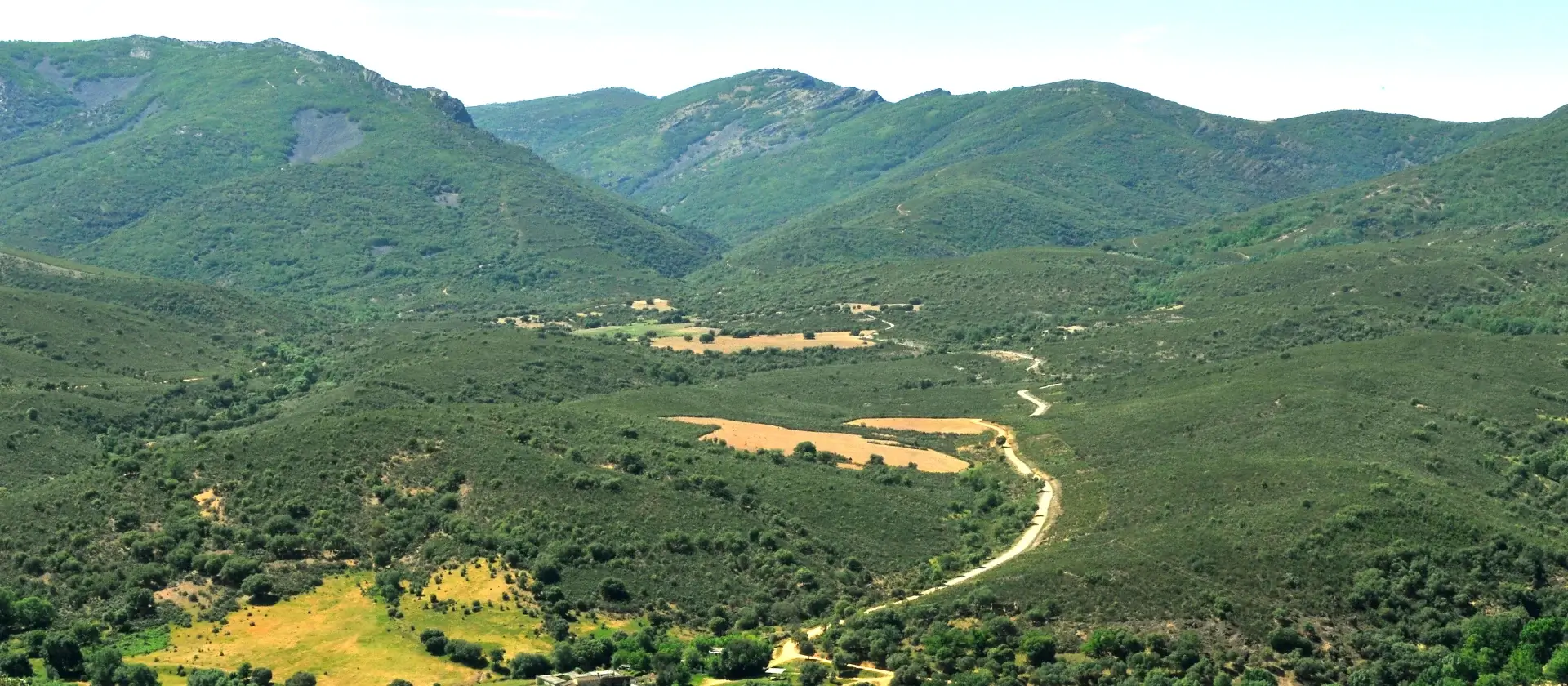 Paisaje montañoso verde con valles y caminos sinuosos, vegetación arbustiva y cielos despejados.