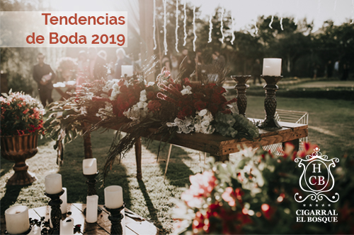 Decoración de boda al aire libre con flores rojas y velas. Texto: "Tendencias de Boda 2019". Logo en la esquina.