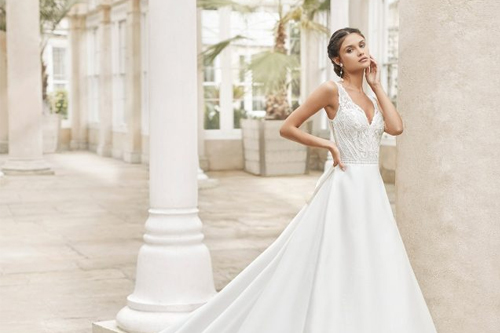 Mujer con vestido de novia blanco en un pasillo elegante, rodeada de columnas y vegetación.