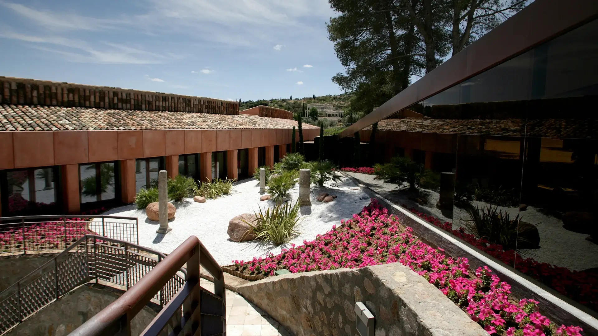 Patio con jardín de grava blanca, plantas y flores rosas. Edificio de techo de tejas y ventanales grandes.
