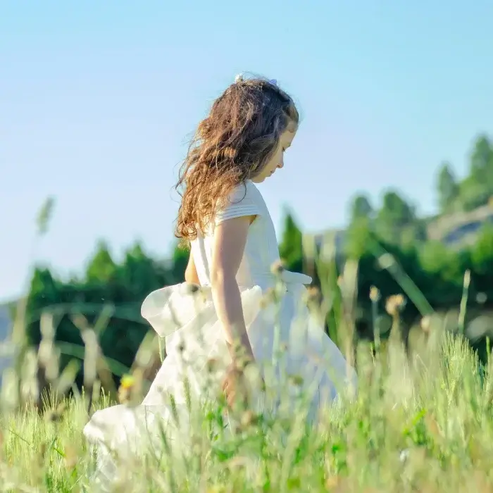 Niña de perfil en un campo verde con un vestido blanco y un gran lazo, mirando hacia abajo, día soleado.