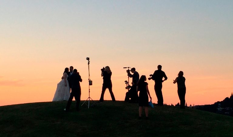 Siluetas de una pareja y fotógrafos en una colina al atardecer, con un cielo anaranjado de fondo.