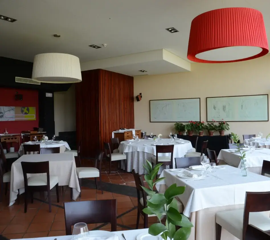 Comedor de restaurante elegante con mesas vestidas y lámparas colgantes grandes, decorado con plantas y cuadros.