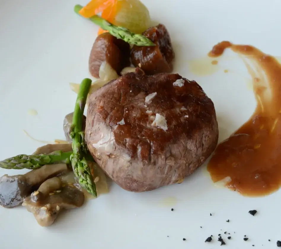 Plato gourmet con filete de carne, espárragos, hongos y aderezo en un plato blanco. Decoración elegante y minimalista.
