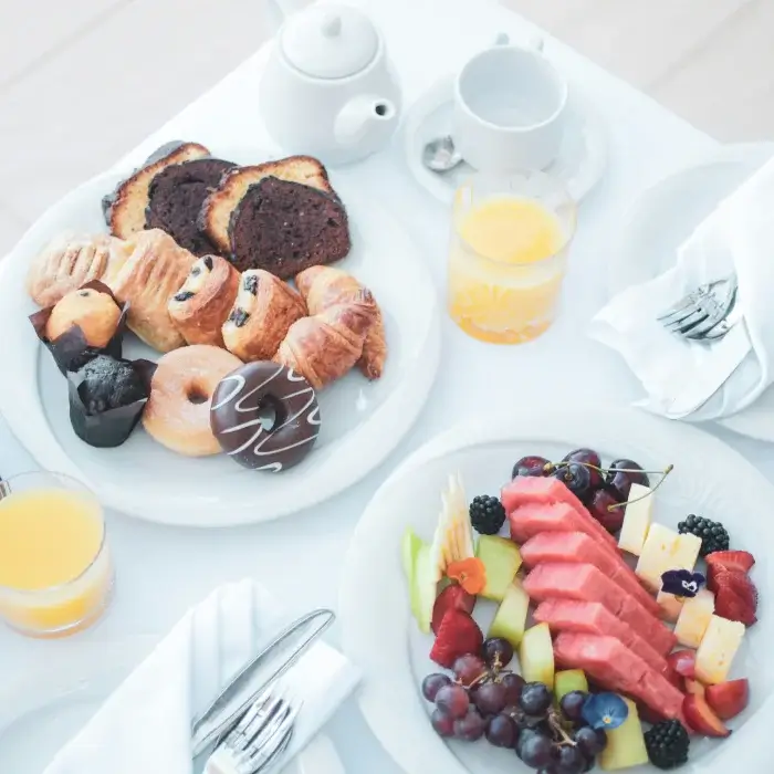 Desayuno con croissants, pastelillos, muffins, fruta variada y jugo de naranja en una mesa blanca.