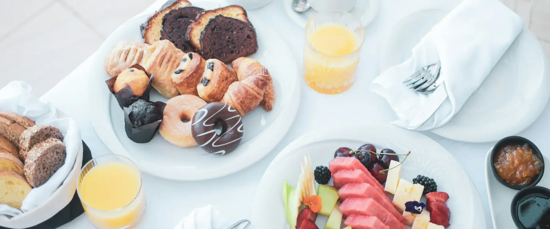 Desayuno variado con panes, pasteles, frutas y jugo de naranja sobre una mesa blanca.