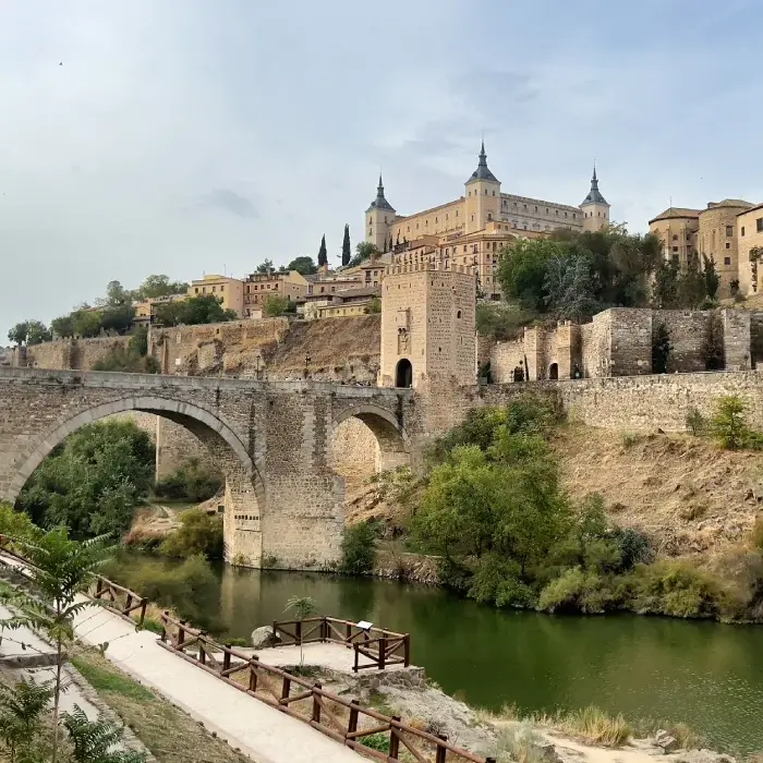 Puente de piedra sobre un río, con una colina en el fondo donde se sitúa un edificio histórico con torres.