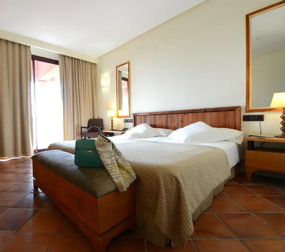 Habitación de hotel con cama amplia, mesitas de noche con lámparas, banco con bolso y bufanda al frente. Cortinas beige.