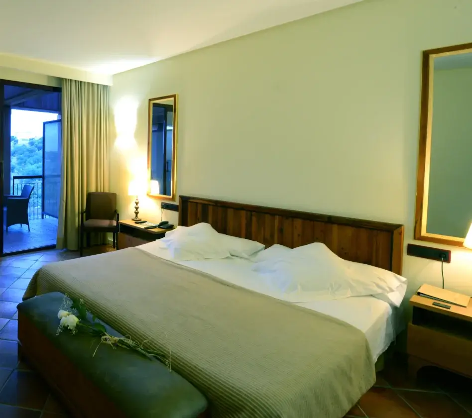 Habitación de hotel con cama doble, lámparas, sillón y balcón con vista exterior. Decoración sencilla y acogedora.