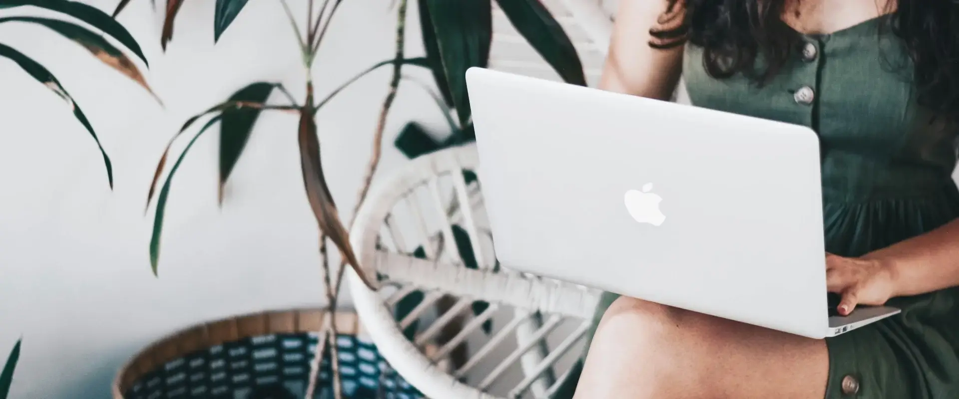 Persona usando una MacBook en una silla de mimbre junto a una planta.
