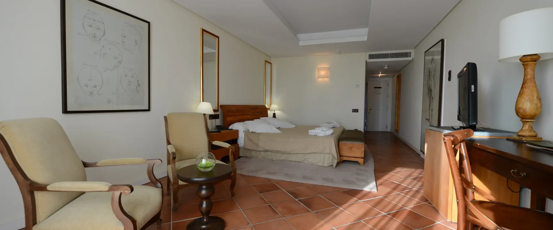 Habitación de hotel con cama doble, sillones, mesa pequeña, lámparas, TV y decoración minimalista.