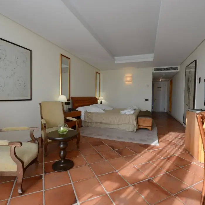 Habitación de hotel luminosa con cama, mesitas, sillones y un sofá. Decoración en tonos claros, con suelo de baldosas.