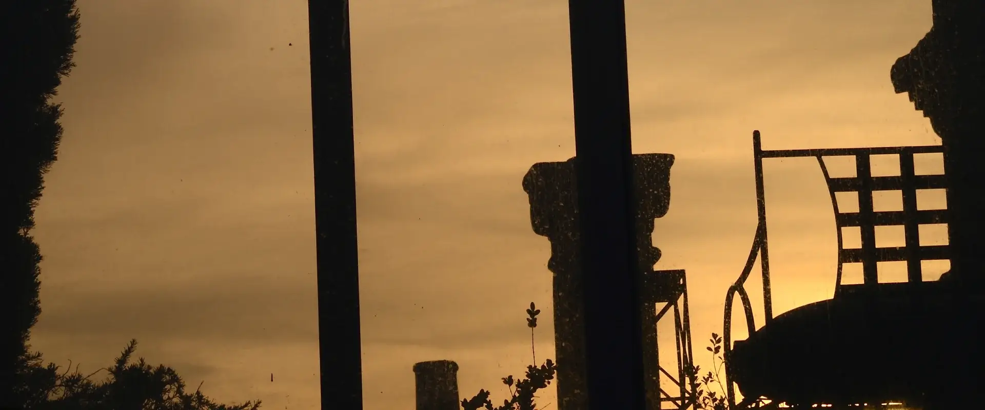 Silueta de una valla y esculturas contra un atardecer naranja, con árboles al fondo y cielo nublado.