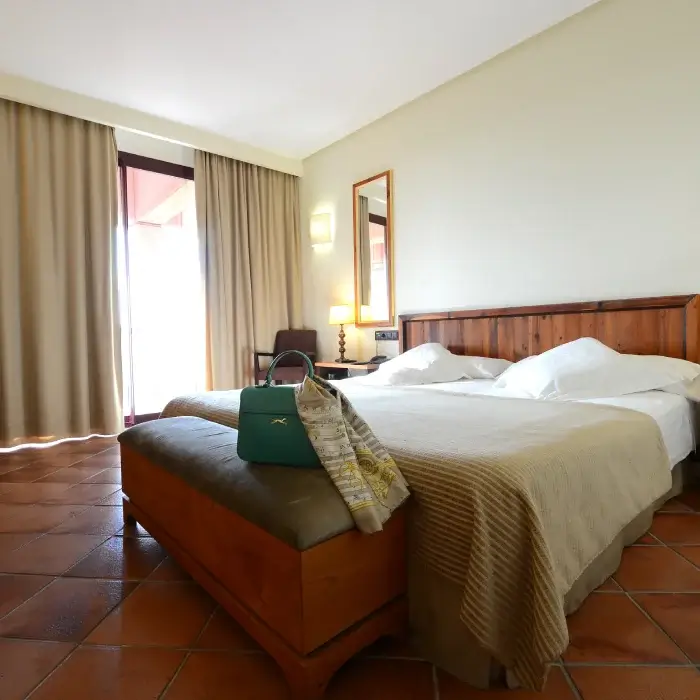 Habitación de hotel con cama doble, luz tenue, cortinas beige, espejo y bolso verde en un banco a los pies de la cama.