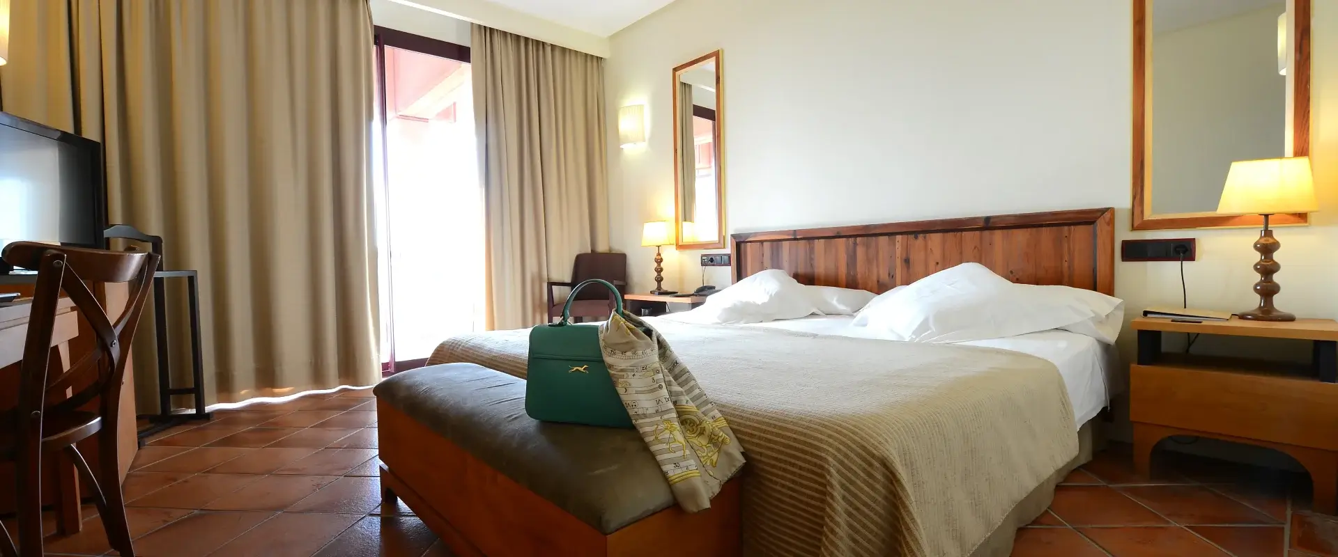 Habitación de hotel con cama doble, lámpara de noche, espejo, cortinas y un bolso verde sobre el banco al pie de la cama.