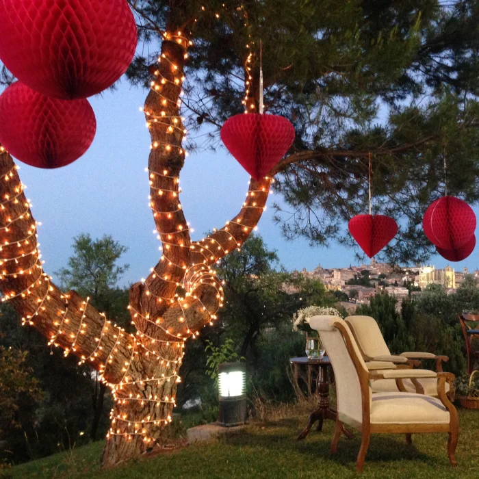 Árbol decorado con luces y farolillos rojos colgantes en un jardín al atardecer, con sillas y vista de la ciudad.