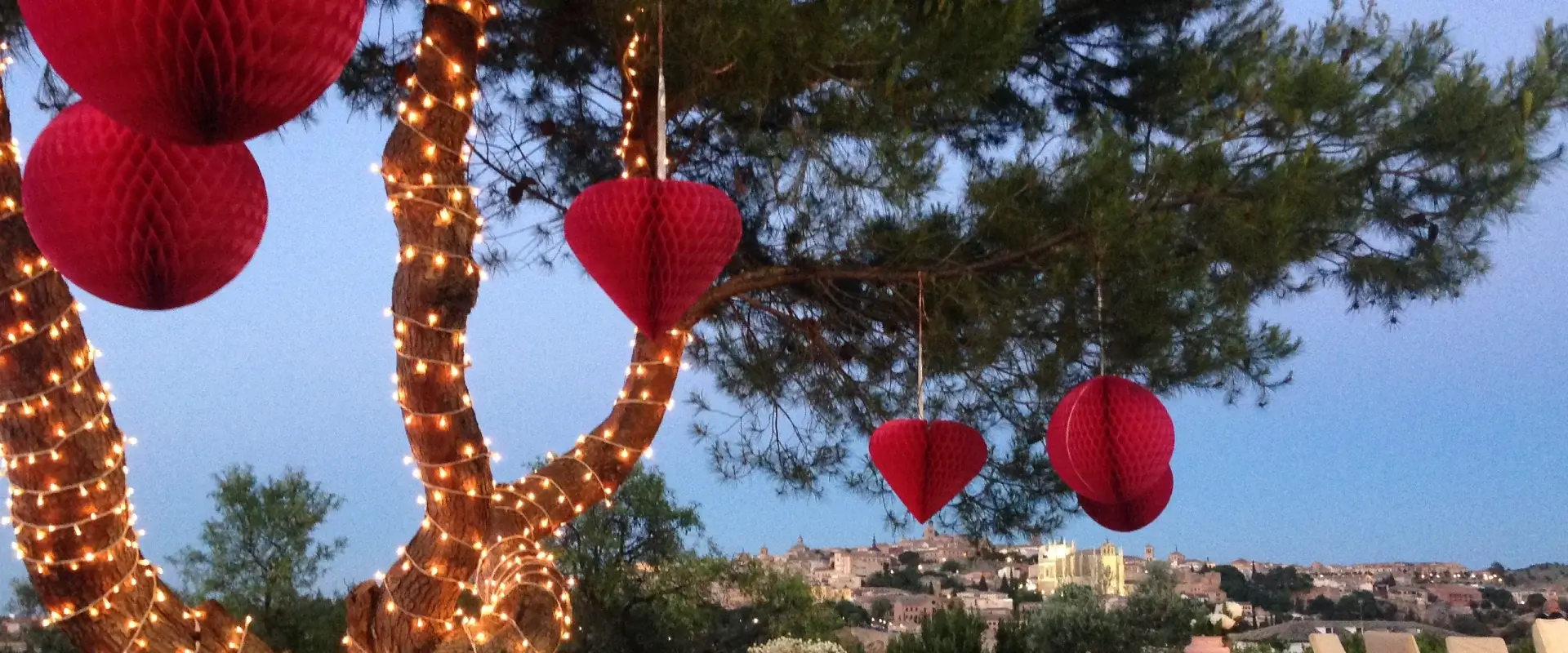 Árbol decorado con luces y adornos rojos de papel colgantes, con vista de una ciudad al fondo al anochecer.