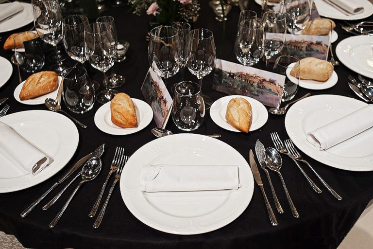 Mesa elegante con platos blancos, cubiertos, copas de vino, panecillos sobre platillos y servilletas enrolladas.