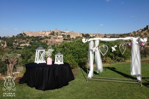 Ceremonia al aire libre con mesa negra con linternas y arco decorado, en un paisaje con ciudad en el fondo.