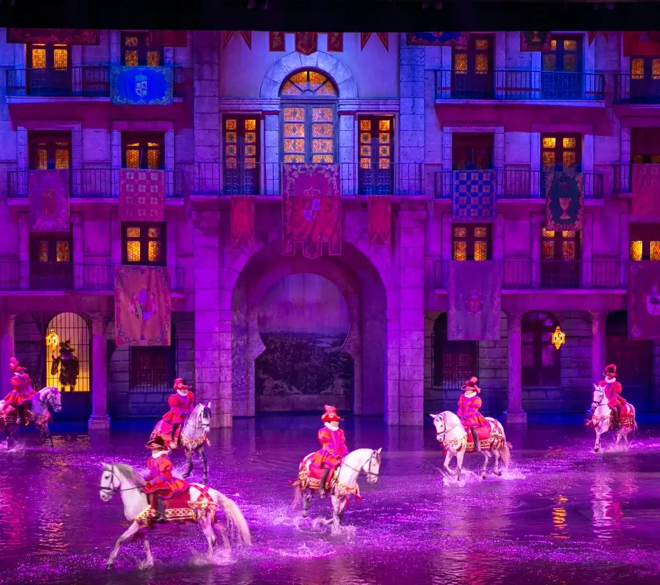Cinco jinetes con trajes rojos en caballos blancos cruzan un escenario acuático frente a un edificio iluminado con decoraciones.