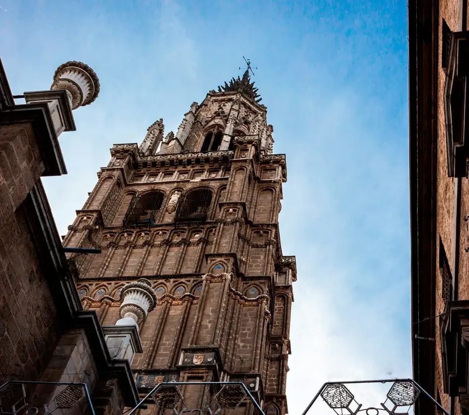 Torre de una catedral gótica vista desde abajo, con cielo azul de fondo. Dos edificios flanquean la escena.