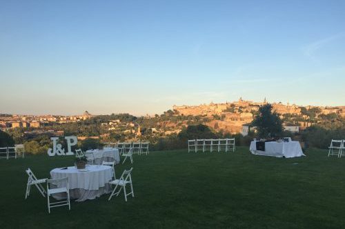 Mesa exterior con decoración de boda J&P y sillas blancas en un jardín, con vista panorámica de una ciudad al fondo.