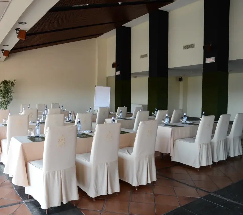 Sala de conferencias con mesas y sillas cubiertas de tela blanca, botellas de agua, pizarra y plantas decorativas.