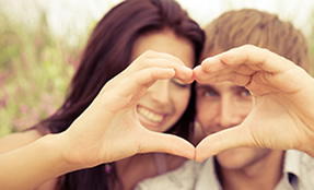 Pareja sonriendo forma un corazón con sus manos, fondo de naturaleza desenfocado.