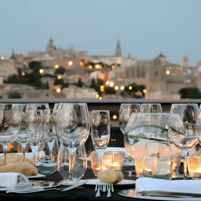 Mesa elegante con copas de vino y velas encendidas, vista difuminada de una ciudad iluminada al fondo.