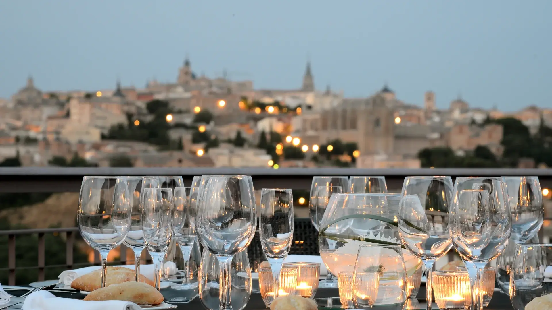 Una mesa elegantemente preparada con copas de cristal, velas y panes, con una ciudad iluminada al fondo al anochecer.