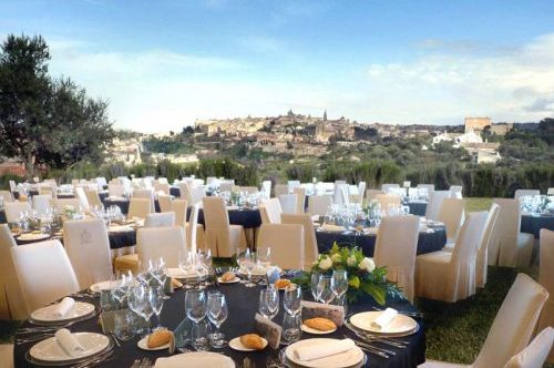 Celebración al aire libre con mesas elegantes, vajilla fina y vista panorámica de una ciudad en el fondo.