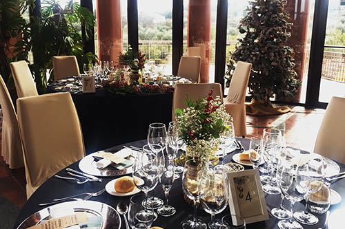 Mesa elegantemente decorada con copas y platos en un salón, junto a una ventana y un árbol navideño adornado.