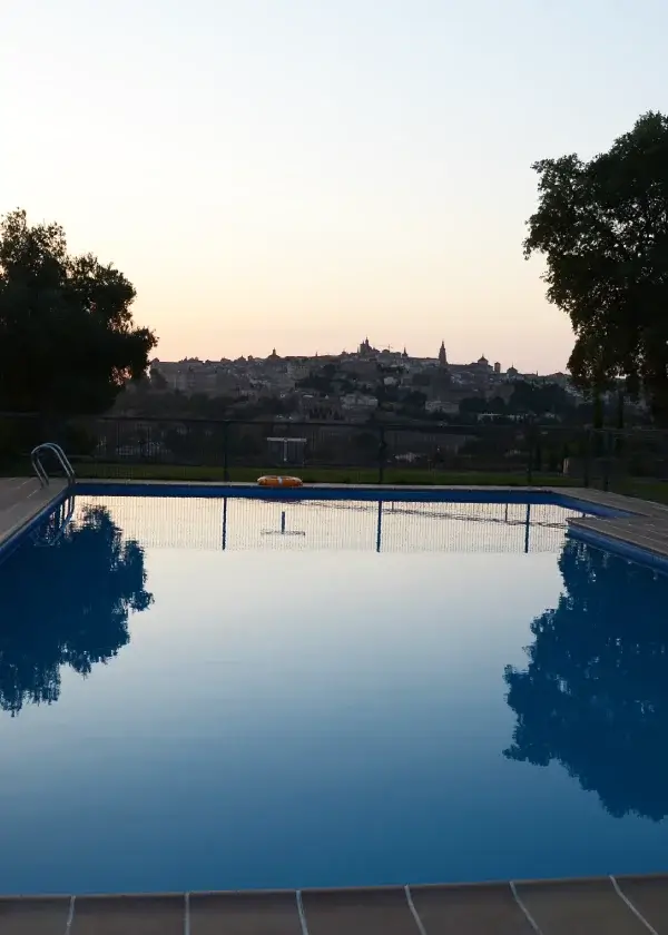 Piscina al atardecer reflejando el cielo, con una ciudad en el horizonte y árboles en los costados.