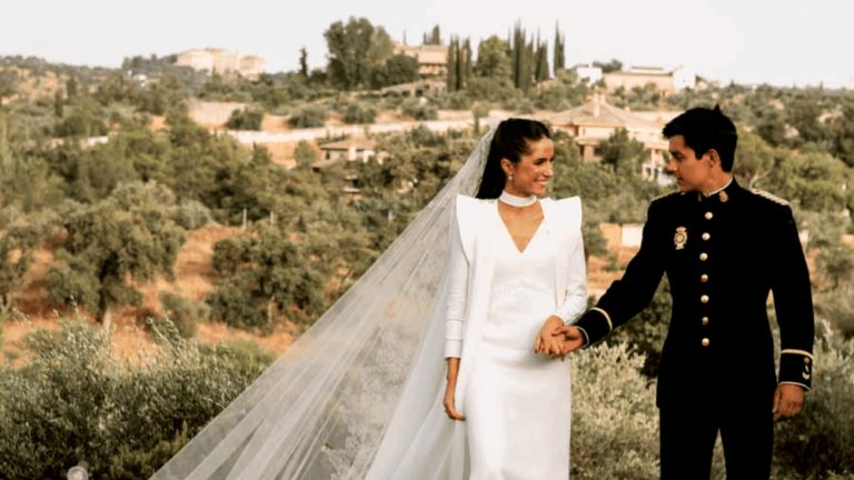 Pareja de novios sonríe tomados de la mano en paisaje rural, ella con vestido blanco y él con uniforme oscuro.