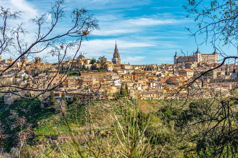 Vista panorámica de una ciudad histórica con edificios antiguos, una catedral y colinas verdes bajo un cielo azul.