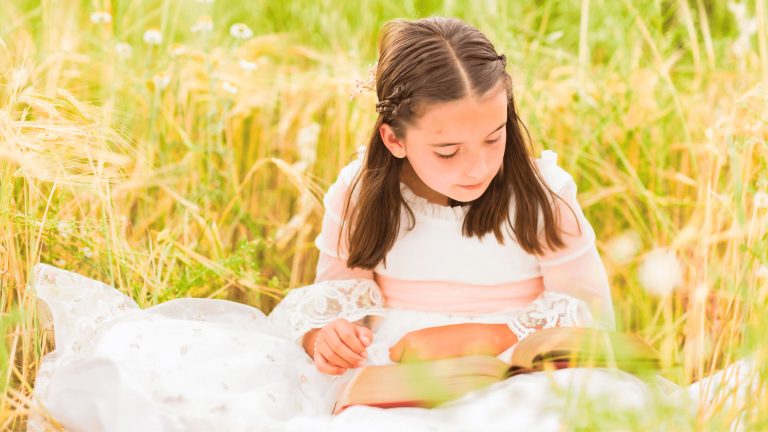 Niña con vestido blanco lee un libro en medio de un campo de césped alto y flores.