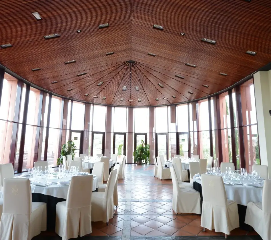 Sala amplia y elegante con mesas redondas, sillas blancas, techo de madera y ventanales grandes.