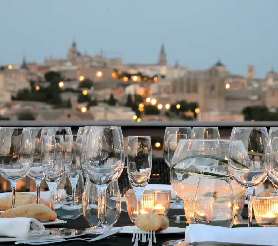 Mesa elegante con copas de cristal y panecillos, al fondo una ciudad iluminada al anochecer.