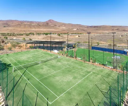 Cancha de tenis al aire libre rodeada de paisaje árido, con una segunda cancha detrás y cerco de malla metálica.