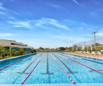 Piscina olímpica al aire libre con carriles, rodeada de sillas y vegetación, bajo un cielo azul despejado.