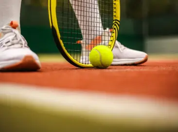Raqueta de tenis junto a una pelota en una cancha de arcilla, con una persona usando zapatillas deportivas blancas.